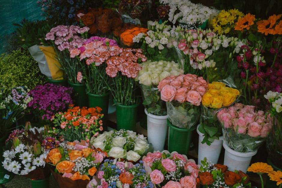An assortment of fresh floral bouquets displayed on a flower shop shelf, featuring various types of roses, carnations, daisies, and other blossoms in shades of pink, white, yellow, orange, and purple.