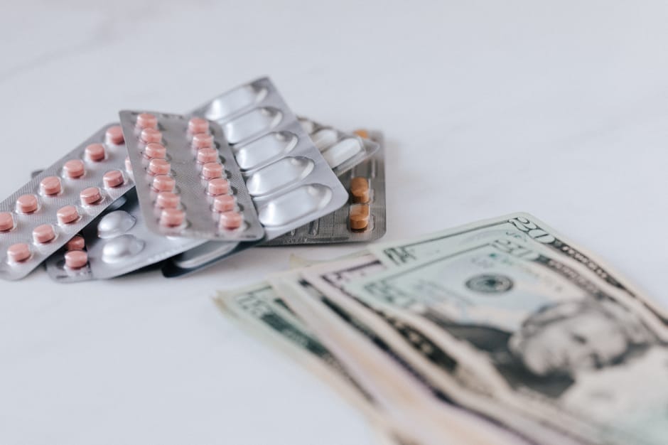 Several blister packs of prescription tablets, including pink and white capsules and silver foil backing, are placed alongside a small stack of US dollar bills on a plain white surface. The arrangemen
