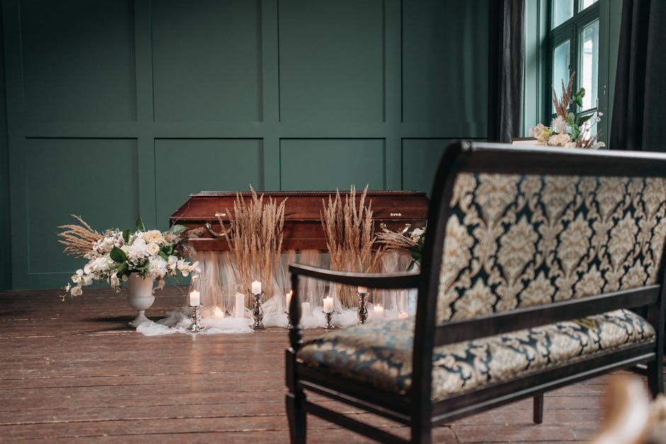 A wooden casket on a dark wooden floor, surrounded by elegant floral arrangements, including white roses, peonies, and greenery in a white vase on the left side, and additional white and pastel flower