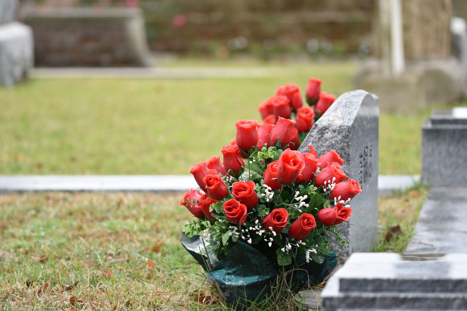 A bouquet of fresh red roses arranged with green foliage and small white filler flowers, wrapped in dark green tissue paper, rests against a grey stone grave marker in a cemetery. The flowers are vibr