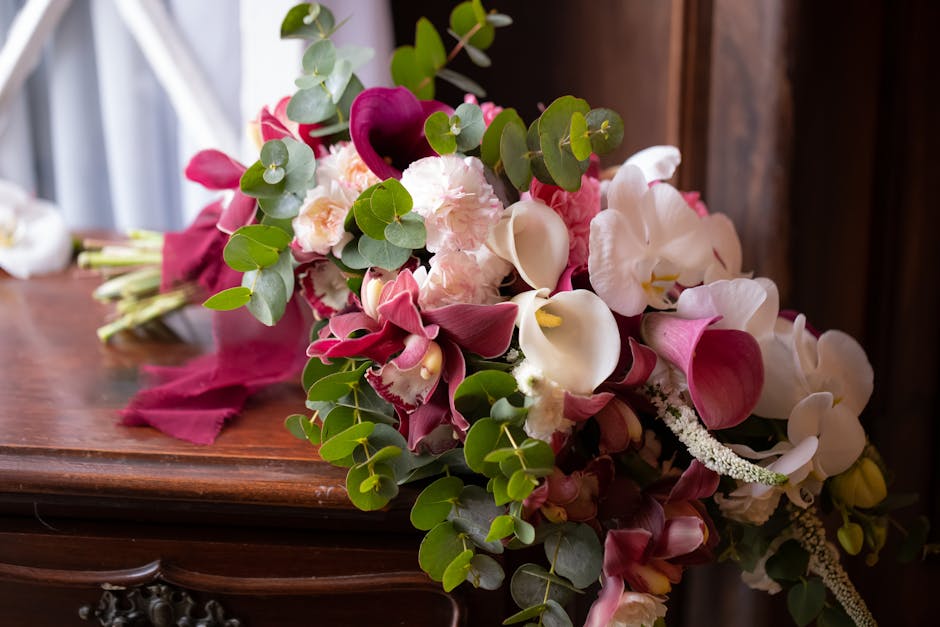 A floral arrangement featuring a cascading bouquet of fresh flowers resting on a polished wooden surface. It includes white and blush pink calla lilies, deep burgundy anthuriums, and soft pink peonies