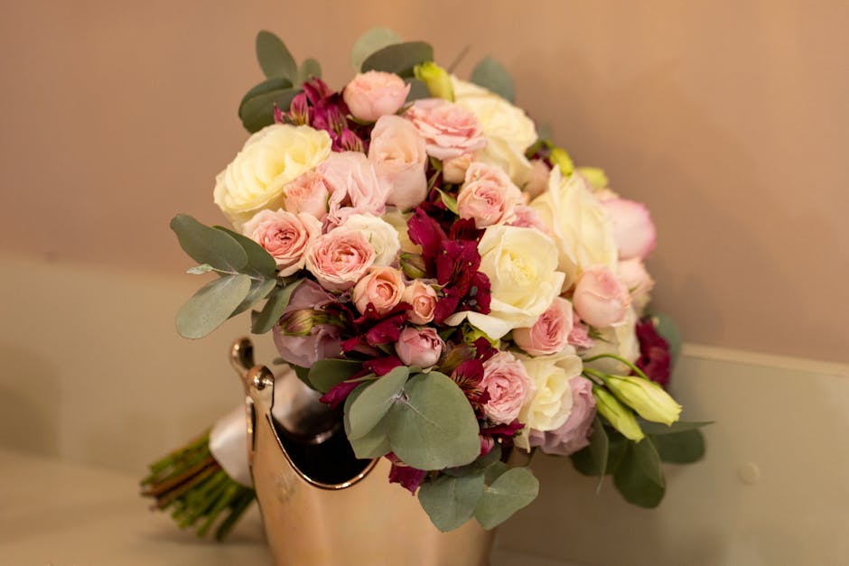 A fresh floral bouquet with a combination of white and soft pink roses, accented by deep burgundy flowers and dusty miller eucalyptus leaves. The arrangement is placed in a metallic gold vase, with th