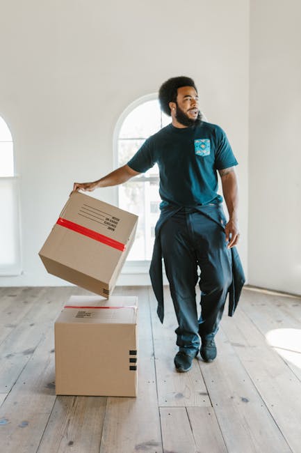 A man with curly dark hair and a beard is standing indoors in front of a large arched window, holding a cardboard box with a red stripe in his right hand, while two additional sealed boxes are stacked