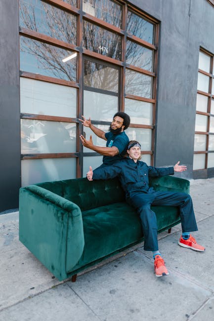 Two smiling men, one standing behind and the other sitting on a plush, emerald green velvet sofa placed on a sidewalk outside a modern building with large wooden-framed windows. The man standing has d